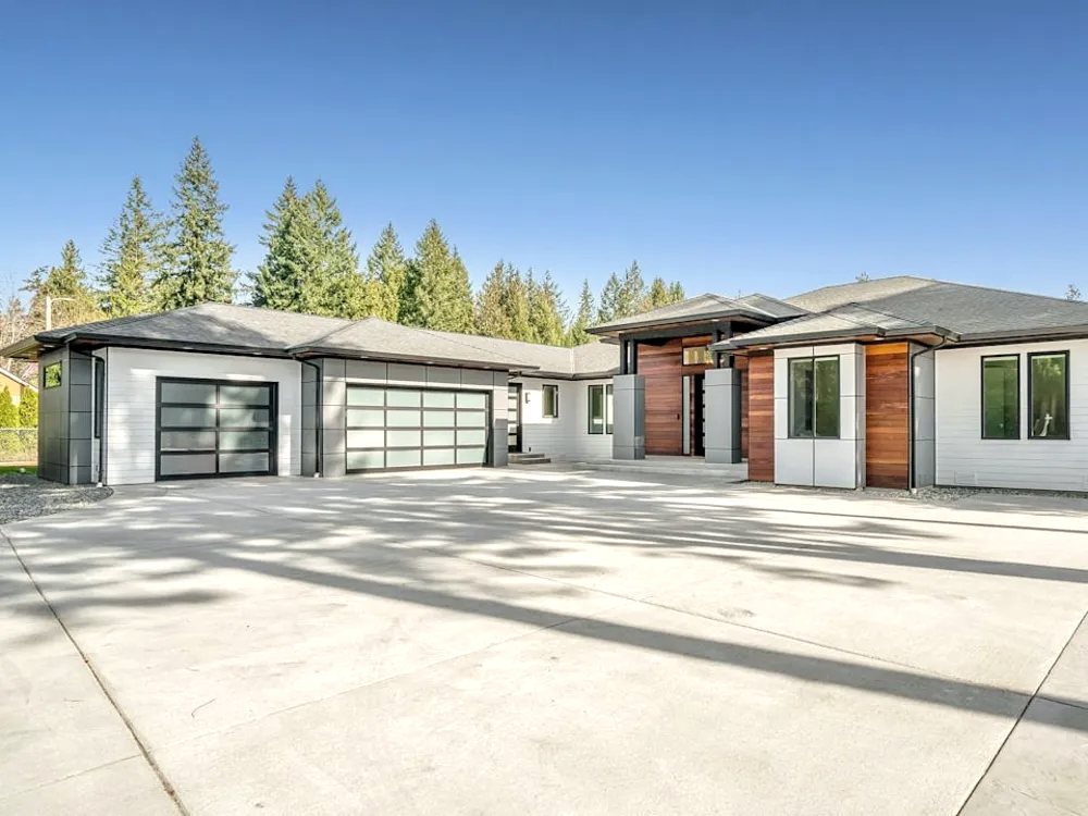 Ranch-style home with a wide concrete driveway and pine trees behind it